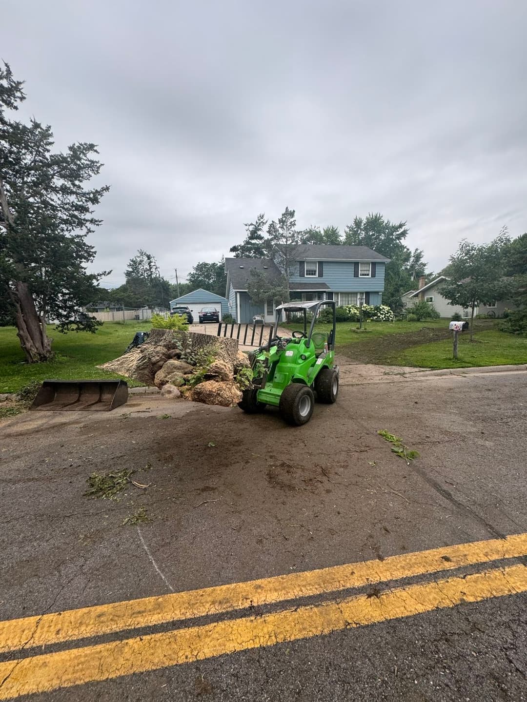 Green compact tractor on a rural road near residential homes with debris in the foreground.